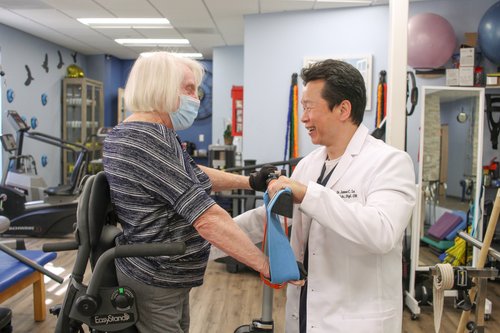 Dr. Lu assisting patient with standing frame and resistance band exercises in rehab gym