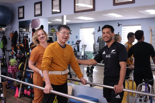 Patient practicing walking between parallel bars during physical therapy with staff encouragement